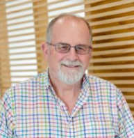 Headshot of a man with grey hair, a beard and glasses in front of a wooden panelled wall.