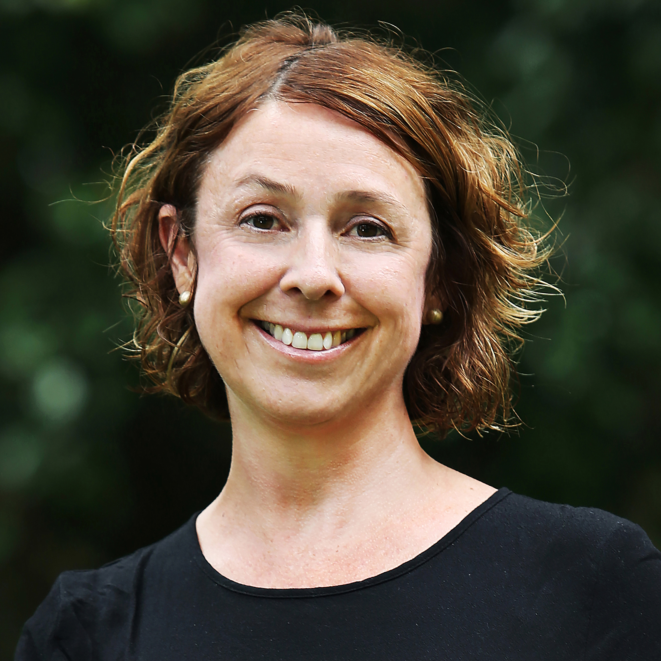 Headshot of a woman with short, brown hair with blurred gardens in the background.