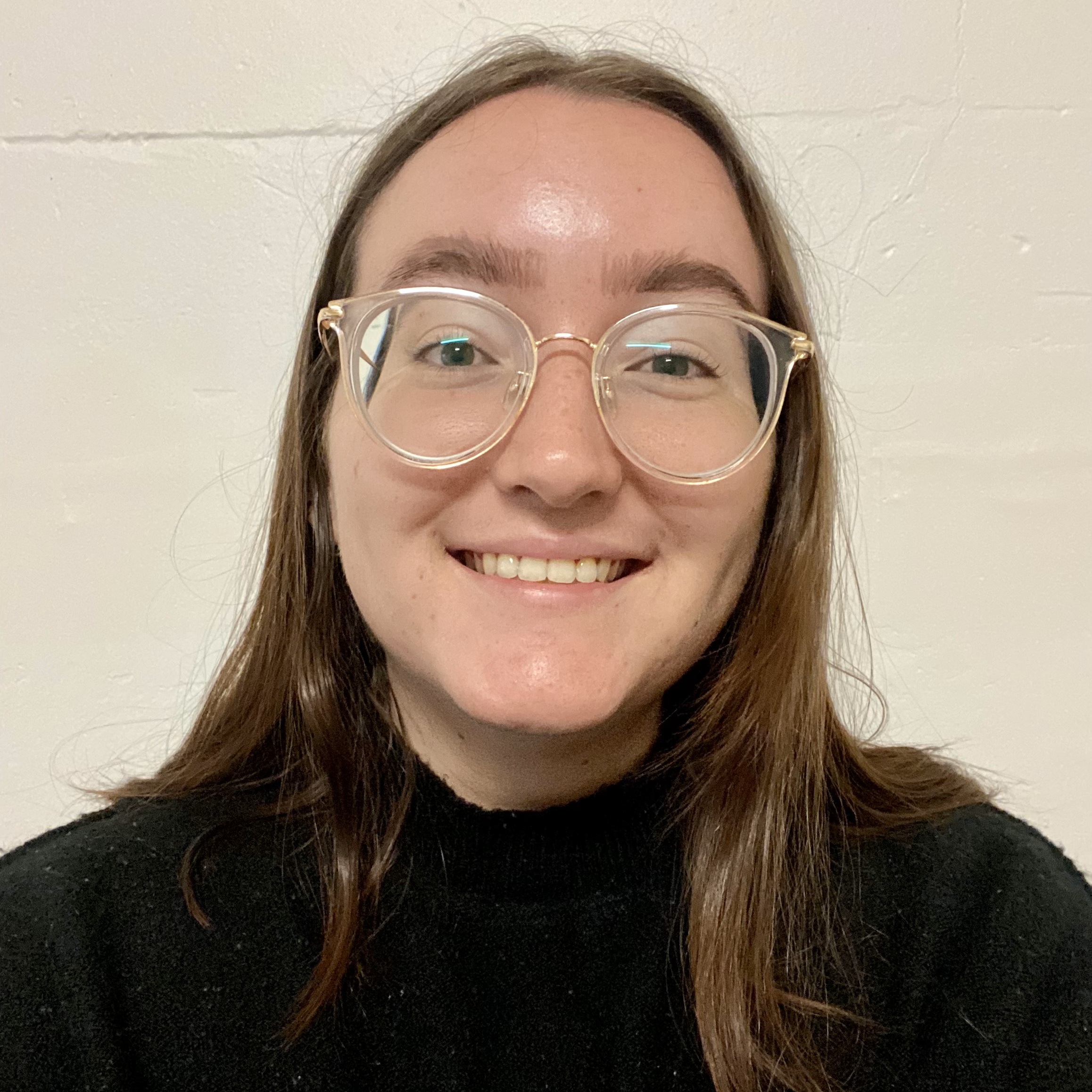 Headshot of a woman with brown hair and glasses in front of a white brick wall.