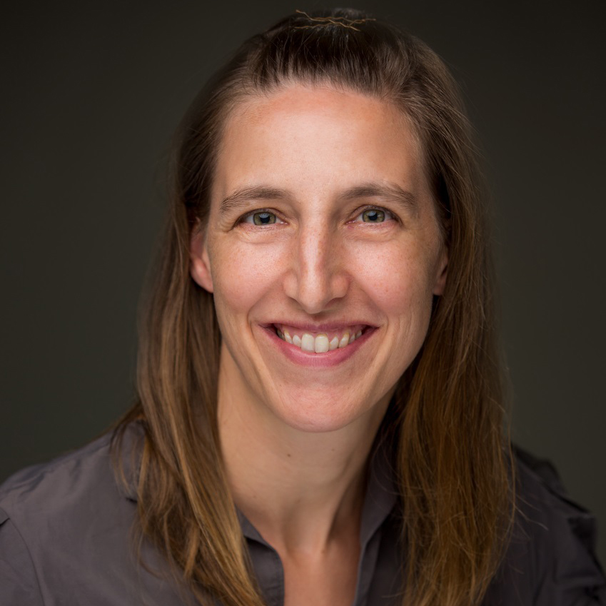 Headshot of a woman with brown hair in front of a grey background.