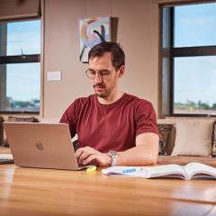A man wearing a maroon shirt and glasses is using a laptop at a desk with an open book and papers on it.